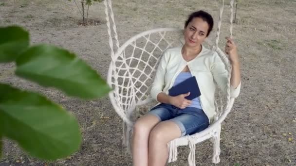 Una mujer dulce y pensativa se balancea en la naturaleza en un columpio blanco con un libro azul en sus manos. Una mujer adulta de nacionalidad oriental en el Parque en un día soleado. Retrato. De cerca. 4K . — Vídeos de Stock