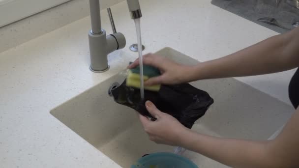 Close-up, womens hands wash a black plate with a yellow sponge under a stream of water in the kitchen sink. Homework. Faceless. Concept. 4K. — Stockvideo