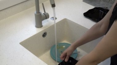 A woman washes dishes, blue and black plates, under a stream of water in the kitchen sink. Closeup. Faceless. Concept. 4K.