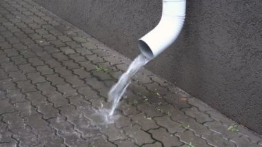 When it rains in the city, water pours from a white drainpipe onto the city sidewalk. Conceptualism. Symbol. Closeup. 4K.