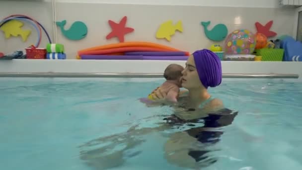 Nado infantil na piscina infantil. O bebé nada cara a cara nos braços da mãe na piscina. A mãe olha para ele e sorri. Fecha a porta. 4K . — Vídeo de Stock