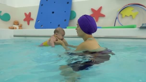 Un bebé recién nacido nada en los brazos de su madre en la piscina infantil. Mamá lo mira y sonríe. Natación infantil. Primer plano. 4K . — Vídeos de Stock