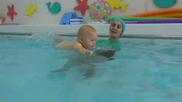 De moeder helpt de baby zwemmen in het kinderbad en ondersteunt hem met haar handen in het water. Ze spelen en lachen. Zwemtraining. Zuigelingen zwemmen. Close-up. — Stockvideo