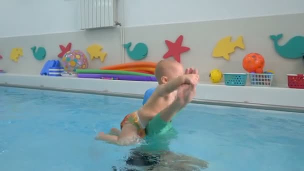 Een vrouwelijke coach doet sportoefeningen met een kind in het water in een kinderbad. Ze omcirkelt het kind in haar armen. Ze spelen en lachen. Zuigelingen zwemmen. Close-up. — Stockvideo