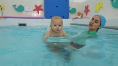 Close-up of a happy toddler learning to swim and swimming with an instructor in a children's pool. He looks around and smiles. Infant swimming. Concept.