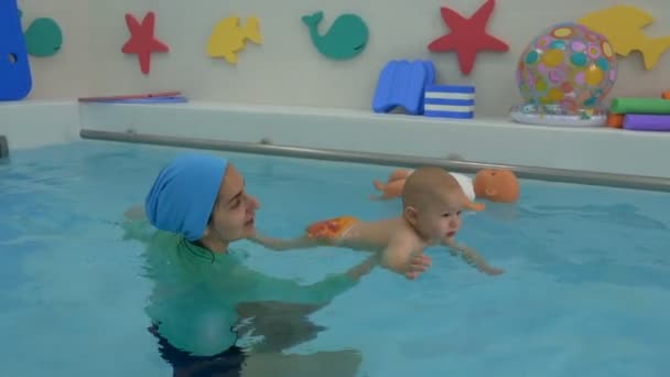 A mãe sustenta o bebê com as mãos, que está flutuando na água na piscina das crianças. O bebê nada para o lado da piscina, olha em volta e sorri. Natação infantil. Fechar. . — Vídeo de Stock