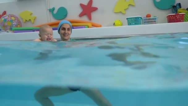 Un bébé pleurant nage dans l'eau dans la piscine des enfants dans les bras de sa mère. La mère apprend à l'enfant à nager et le soutient. Nage infantile. Plan divisé. Sous l'eau et à la surface . — Video