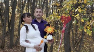 A pregnant couple in love hugs and kisses in the autumn forest. A man and a pregnant young girl pose against a background of a toy heart and the sun, which are hung on the branches of a tree. Closeup.