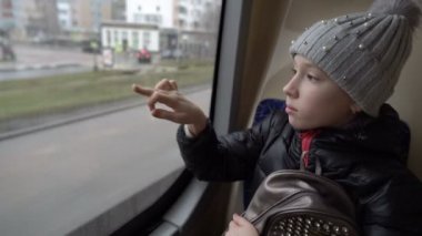 A sad little girl is riding a bus, looking out the window and drawing on the dirty glass, sitting in the back seat. View from inside the bus. Blurred background. Close-up. 4K.