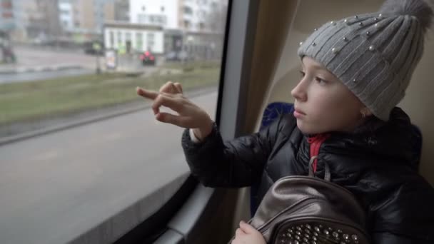 Una niña triste está montando un autobús, mirando por la ventana y dibujando en el vidrio sucio, sentada en el asiento trasero. Vista desde el interior del autobús. Fondo borroso. Primer plano. 4K . — Vídeo de stock