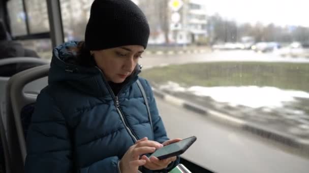 Een volwassen meisje zit bij het raam in een bus door de stad te reizen en kijkt naar iets op haar smartphone. Huizen, auto 's en mensen flitsen voorbij het raam. Een close-up. Wazige achtergrond. — Stockvideo