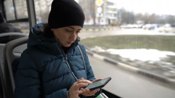 Una chica adulta está sentada cerca de la ventana en un autobús que viaja por la ciudad y está mirando algo en su teléfono inteligente. Casas, coches, y la gente pasa por la ventana. Primer plano. Fondo borroso . — Vídeo de stock