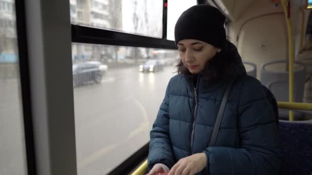 Een volwassen Oosters meisje staat in de bus bij het raam en haalt een creditcard uit haar portemonnee om de rit te betalen. Auto 's en huizen flitsen voorbij het raam. Een close-up. Wazige achtergrond. 4k. — Stockvideo