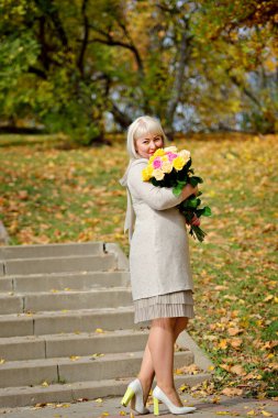 Orta yaşlı sarışın bir kadın elinde bir buket sarı gülle sonbahar ağaçlarına karşı durup kameraya bakar. Moda portresi. Dikey görünüm.