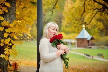Ellerinde kırmızı güller olan tombul, orta yaşlı bir sarışın güzel bir kırsal alana ve küçük ahşap bir kiliseye karşı poz veriyor. Yakın plan. Yatay görünüm.