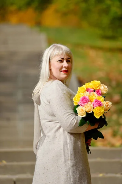 Duża blondynka stoi i pozuje z bukietem żółtych i różowych róż w parku, na zewnątrz, na tle drzew i patrzy w aparat przez ramię. Portret mody. — Zdjęcie stockowe