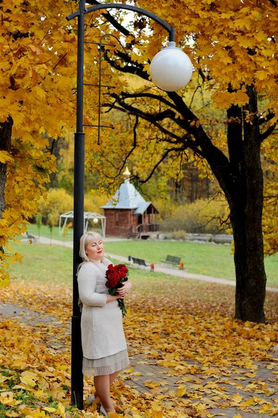 A middle-aged blonde woman stands leaning against a lantern in a city Park against a background of yellow autumn trees with red roses in her hands. She looks into the distance and smiles. — Stock Photo, Image