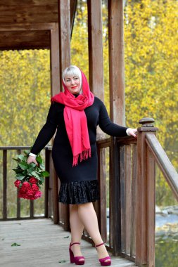 Kırmızı başörtülü, orta yaşlı, çekici bir kadın siyah elbiseli, eski bir ahşap evin verandasında poz veriyor. Gülümsüyor ve elinde bir demet kırmızı gülle uzaklara bakıyor..