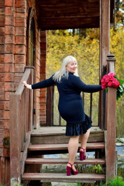 Tombul, oyuncu, orta yaşlı bir sarışın eski ahşap bir evin merdivenlerinde siyah bir elbiseyle dikiliyor, gülümsüyor ve kameraya bakıyor. Dikey görünüm.