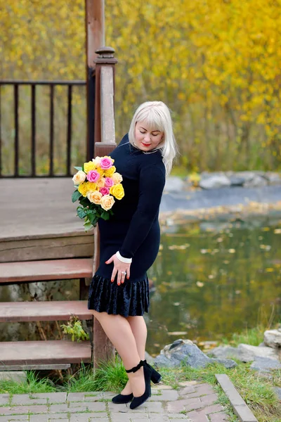Miła, duża kobieta stoi w czarnej sukience i patrzy na swoje czarne buty. Pozuje na zewnątrz z bukietem żółtych i różowych róż w rękach. Portret mody. Widok pionowy. — Zdjęcie stockowe