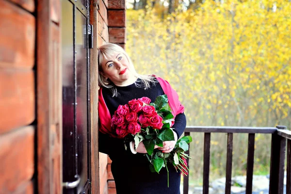 Una mujer de mediana edad de ensueño, de gran tamaño, con el pelo blanco, posa al aire libre cerca de una casa de madera con rosas rojas en las manos sobre un fondo de follaje amarillo. Primer plano. Retrato . —  Fotos de Stock