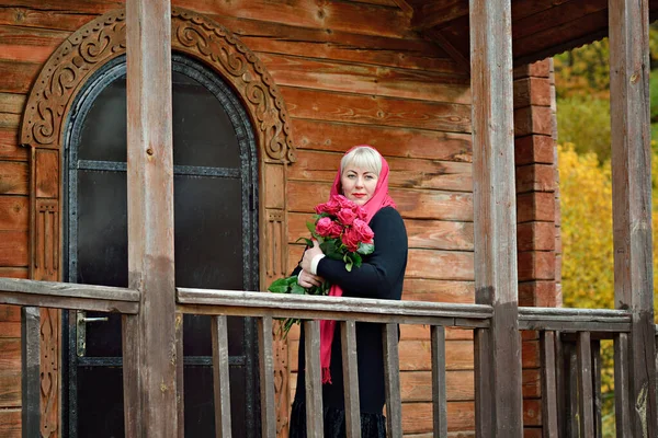 Una mujer grande, triste, de mediana edad, se para en el porche de una casa de madera con rosas rojas en las manos, con un pañuelo rojo y un vestido negro, y mira pensativamente a la cámara . —  Fotos de Stock