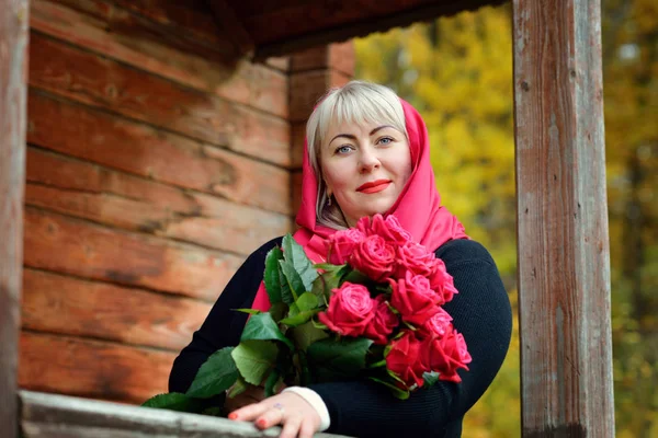 Portret uroczej blondynki w średnim wieku w czerwonym szaliku z czerwonymi różami w rękach. Pozuje w naturze na ganku starego drewnianego domu w czarnej sukience. Uśmiecha się i patrzy w kamerę.. — Zdjęcie stockowe