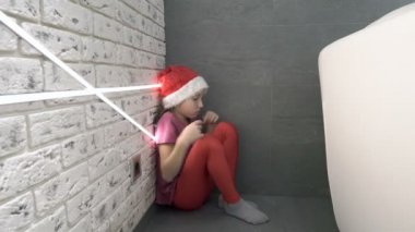 Noel Baba şapkalı gücenmiş küçük bir kız köşedeki evin zemininde oturmuş dizlerine sarılıyor ve gözyaşlarını siliyor. - Yakın çekim. Kavram. 4k.