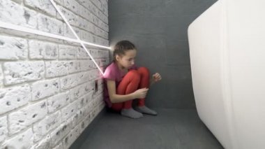 Üzgün küçük bir kız odanın bir köşesinde koltuğun arkasında oturuyor, dizlerine sarılıyor ve gözyaşlarını siliyor. Kızgınlık, depresyon. Kavram. - Yakın çekim. 4k.