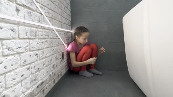 Seorang gadis kecil yang sedih sedang duduk di sudut ruangan di lantai belakang sofa, memeluk lututnya dan menyeka air mata. Kebencian, depresi. Konsep. Penutup. 4K . — Stok Video