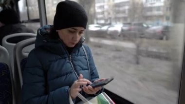 Sıcak giysili yetişkin bir kız akıllı telefonunun sayfalarını çevirir şehirde dolaşan bir otobüsün camında oturur. Otobüsün kabininden bir görüntü. Yakın çekim..