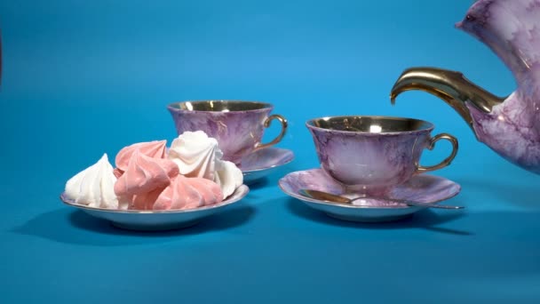 Černý čaj se nalévá do krásných růžových porcelánových pohárků na modrém pozadí. Vedle něj je talíř s marshmallows. Zátiší na modrém pozadí. Koncept. Detailní záběr. 4k. — Stock video