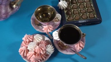 Çay güzel antika porselen bardaklara dökülür. Yanında da bir kutu çikolata ve marşmelovlu pembe bir fincan tabağı var. Mavi arka planda çay hala hayat dolu. Yukarıdan manzara. - Yakın çekim. 4k.
