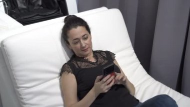 Yetişkin bir doğulu kadın, bir odadaki beyaz bir kanepede siyah giysiler içinde yatarken akıllı telefon kullanır. İnternetten alışveriş. Kavram. - Yakın çekim. 4K.