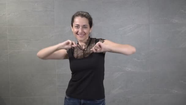 Una dulce mujer oriental de mediana edad baila felizmente en casa frente a la cámara contra una pared gris. Se ríe, posa y mira a la cámara. Retrato de moda. 4K . — Vídeos de Stock