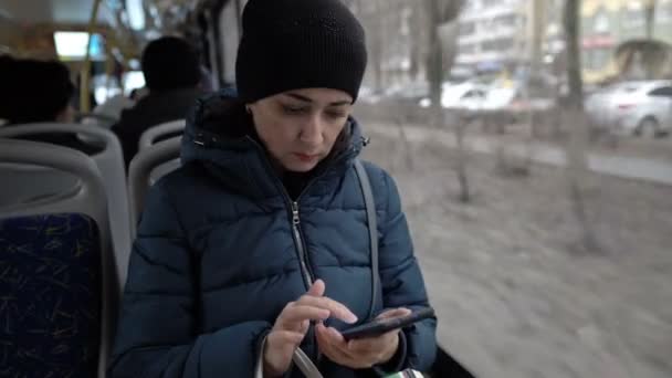 Close-up, una chica adulta con un teléfono en sus manos está sentada en la ventana de un autobús que pasa por la ciudad con ropa de abrigo y mirando algo en la pantalla de un sfartphone . — Vídeos de Stock