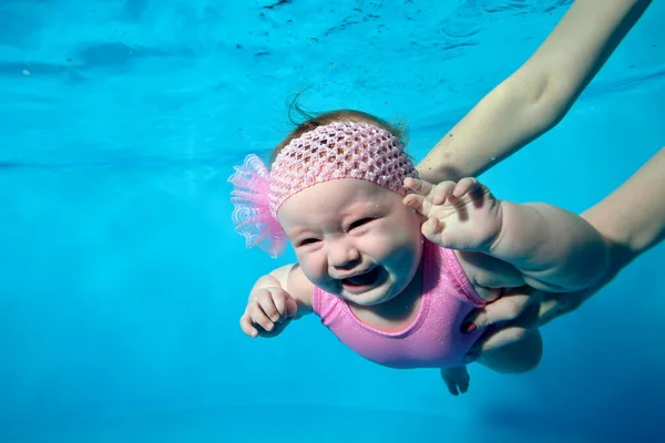 A crying little girl swims under the water in a red swimsuit, and the hands of an adult support her. Fashion portrait. Horizontal orientation. — Stock Photo, Image