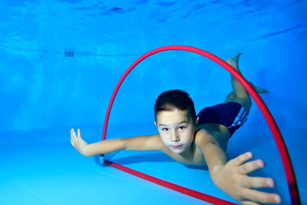 Un niño pequeño y lindo se sumerge hasta el fondo en una piscina para niños y nada a través de un aro rojo sobre un fondo azul. El chico mira a la cámara. Foto digital. Orientación horizontal . — Foto de Stock