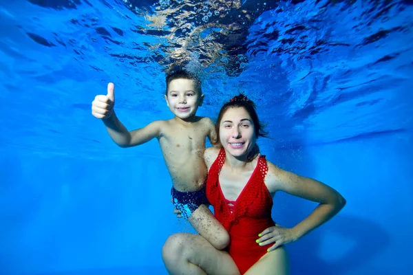 Una madre feliz y su hijo pequeño posan para la cámara bajo el agua en una piscina sobre un fondo azul. Mamá con un traje de baño de punto rojo mira a la cámara. Sonríen. Primer plano. Fotografía digital . — Foto de Stock