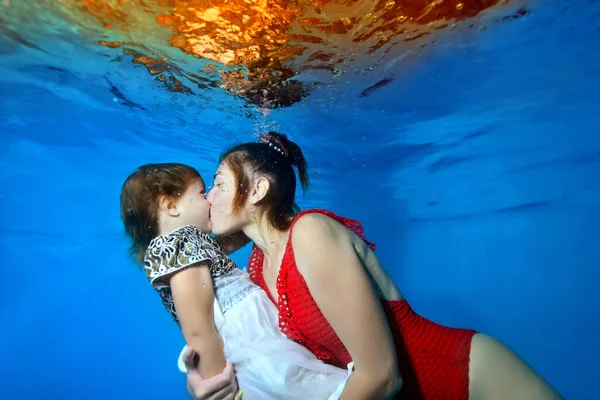 Mutlu bir yetişkin anne ve küçük kızı, baloncuklarla çevrili mavi bir arka planda suyun altında oynaşıp öpüşüyorlar. Kırmızı örülü mayo giymiş bir anne, elbise giymiş bir kız. Yakın plan.. — Stok fotoğraf