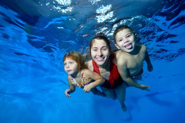 Família de esportes: uma mãe sorridente e seus dois filhos pequenos nadam debaixo d 'água na piscina. Eles abraçam, sorriem e posam para a câmera em um fundo azul. Fecha. Foto digital . — Fotografia de Stock