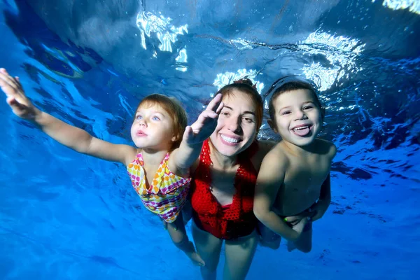 Niños felices, un niño y una niña, junto con su madre nadan y juegan bajo el agua en la piscina. Sonríen provocativamente y posan para la cámara sobre un fondo azul. Fotografía digital . — Foto de Stock