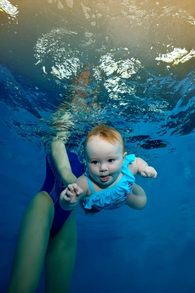 A newborn baby dives under the water in a childrens pool on a blue background. Mother teaches the baby to swim. Fashion portrait. Digital photo. Vertical orientation. — Stock Photo, Image