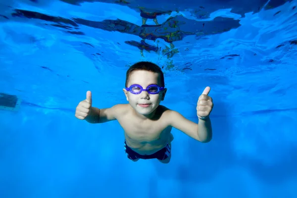 Un niño deportivo posa para la cámara bajo el agua en una piscina infantil con gafas de baño. Él mira a la cámara y da un pulgar hacia arriba. Primer plano. Concepto. Foto digital. Vista horizontal . — Foto de Stock