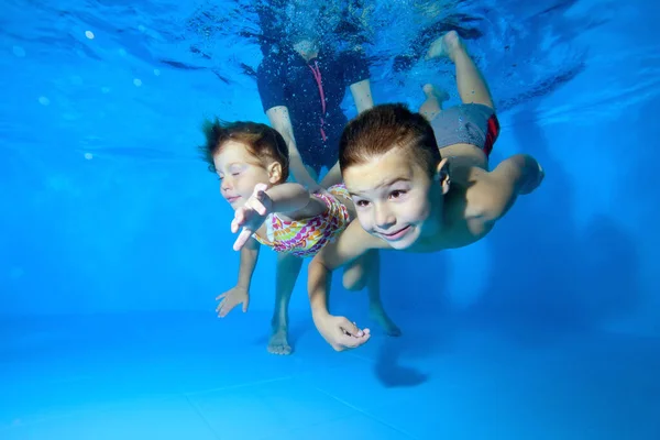 Dos niños, un niño pequeño y una niña, se dedican a nadar en una piscina infantil. Juegan y bucean bajo el agua con los ojos abiertos, y una instructora les ayuda. De cerca. Concepto . — Foto de Stock