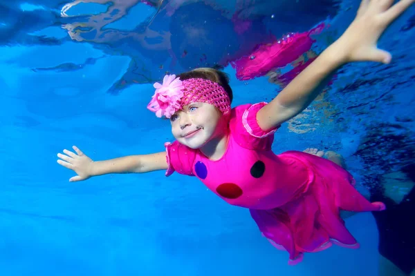 Una linda niña con un hermoso vestido rojo está nadando y sonriendo bajo el agua en una piscina para niños con los brazos extendidos sobre un fondo azul. Primer plano. Concepto. Foto digital. Vista inferior . — Foto de Stock