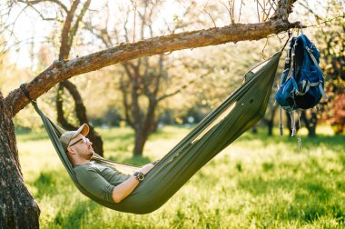 Mutlu traveler sakallı hipster yaz güneşli gün tatil turuncu çiçek açan bahçede elma ağacı üzerinde yatan yeşil şapkalı adam. Hafta sonu ve tatil. Turizm. Boş zaman. Rüya. Koku havada.
