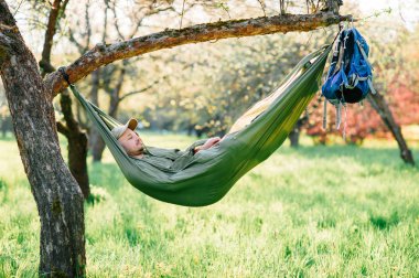 Mutlu traveler sakallı hipster yaz güneşli gün tatil turuncu çiçek açan bahçede elma ağacı üzerinde yatan yeşil şapkalı adam. Hafta sonu ve tatil. Turizm. Boş zaman. Rüya. Koku havada.