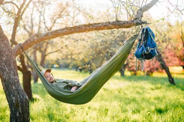 Mutlu traveler sakallı hipster yaz güneşli gün tatil turuncu çiçek açan bahçede elma ağacı üzerinde yatan yeşil şapkalı adam. Hafta sonu ve tatil. Turizm. Boş zaman. Rüya. Koku havada.
