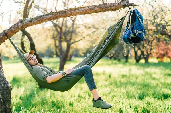 Happy traveler bearded hipster man in green hat lying in hummock on apple tree in blooming garden in summer sunny day on vacation. Weekend and holiday. Tourism. Leisure. Dreaming. Fragrance in air. — Stock Photo, Image